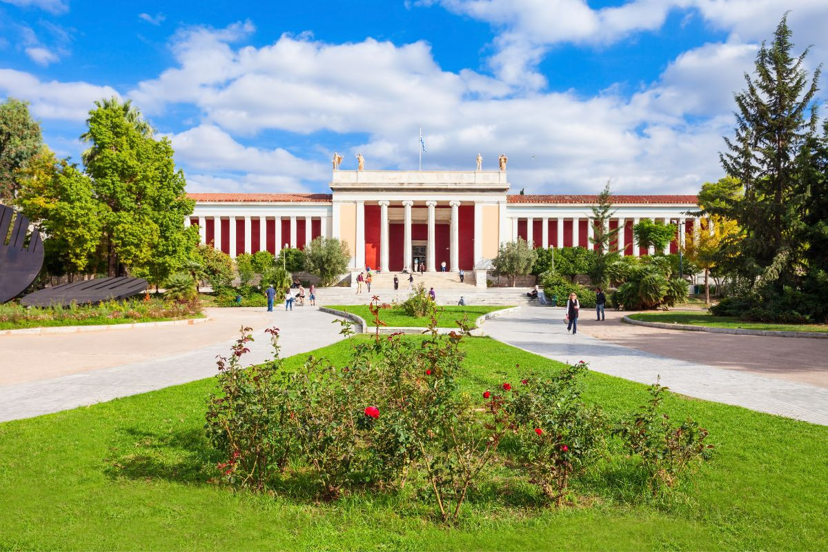 The image shows the National Archaeological Museum of Athens, a neoclassical building with red and white columns. A garden with green grass and red flowers is in the foreground, and several people are walking towards the entrance. The sky is partly cloudy with blue patches_top 10 places to visit in Athens